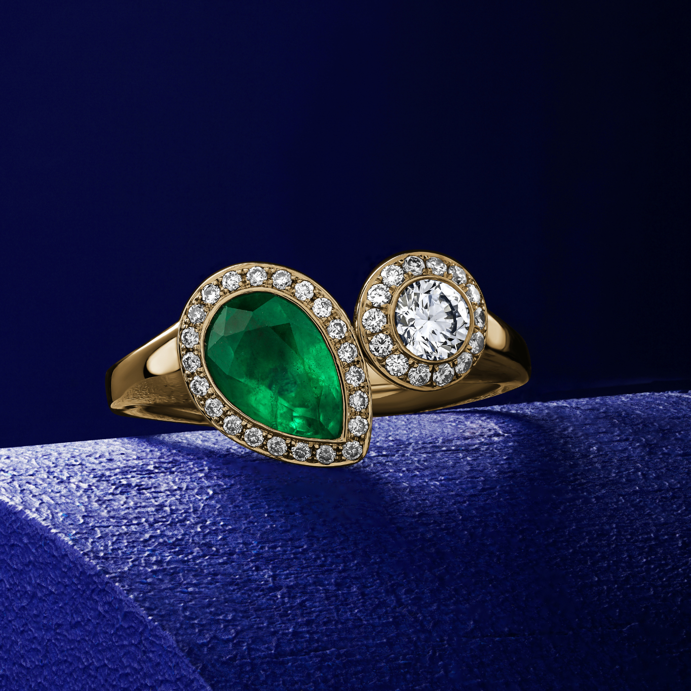 Toi et moi ring featuring a pear-shaped emerald and a smaller round diamond surrounded by a halo of smaller diamond accents designed by carati jewelry in montreal canada in 18 k 14 k diamonds IN blue backround
