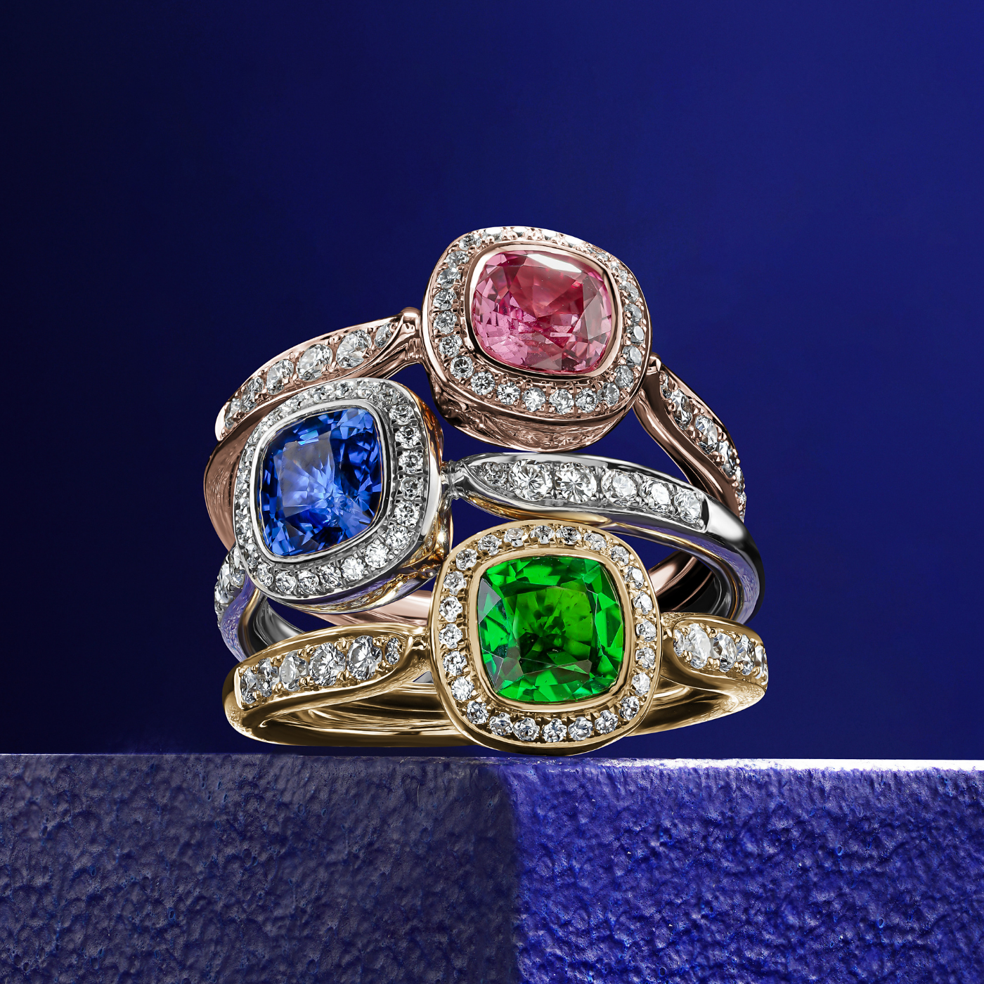 Royal harmony rings with pink, blue and tsavorite cushion surrounded by pave diamonds and on the shank designed by carati jewelry in montreal canada 14k 18 k in blue backround