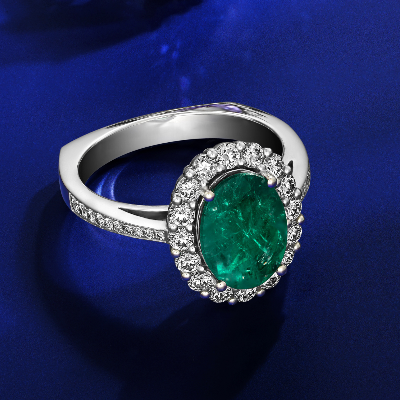 An oval emerald engagement ring with a halo of round diamonds around it and on the band, set in a white gold metal designed by carati jewelry montreal canada in 18k 14 k in blue backround