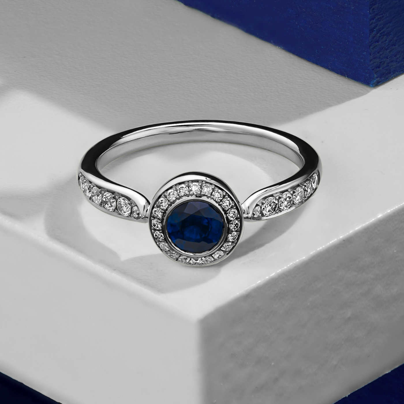 Royal harmony ring with round royal blue sapphireb surrounded by pave diamonds and on the shank designed by carati jewelry in montreal canada 18k 14 k in zoom and white backround