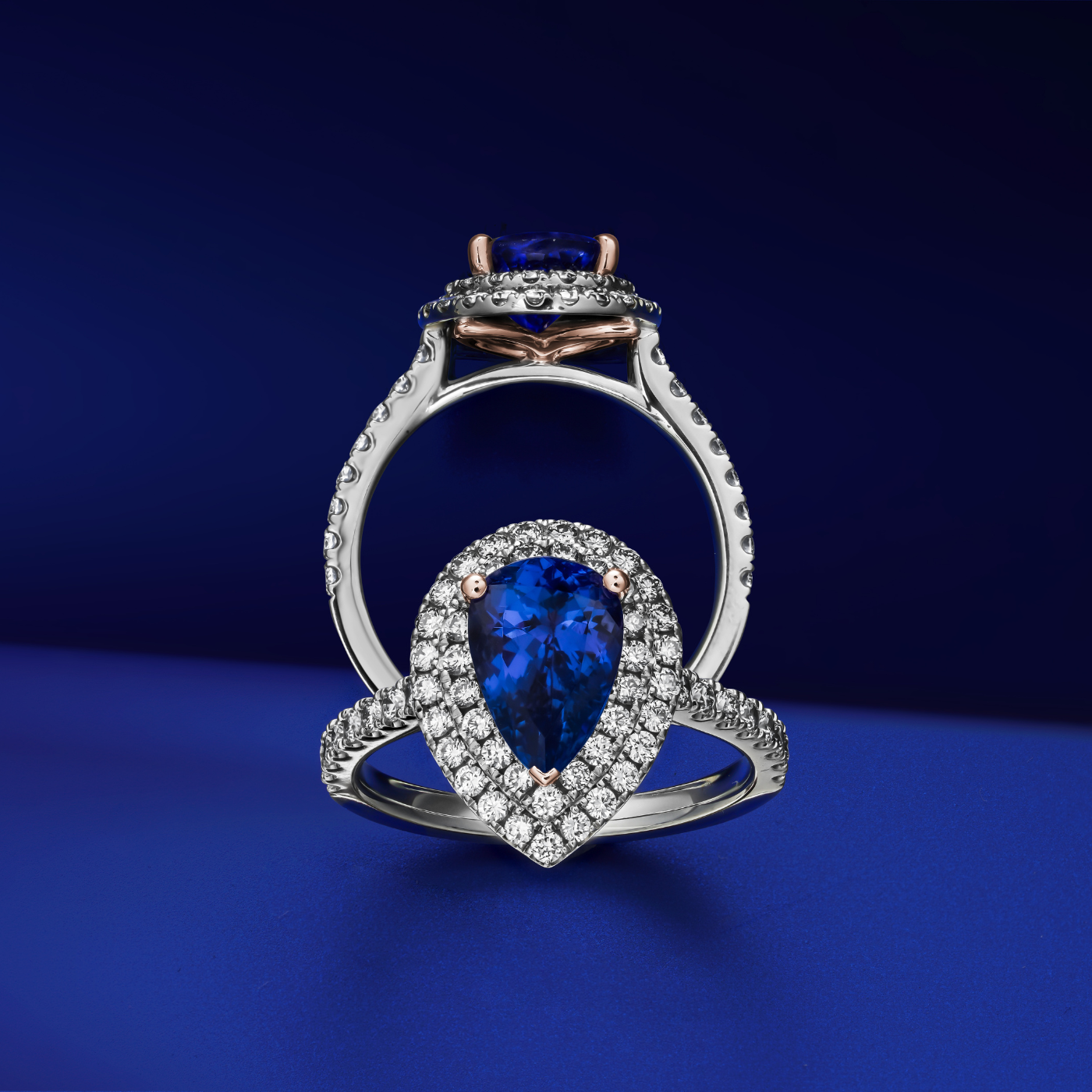 A two row two tone halo ring centered with a pear-shaped tanzanite stone and surrounded by smaller diamonds set in a silver or white gold band from carati jewelry montreal canada in a blue backroud found on instagram caratiofficial