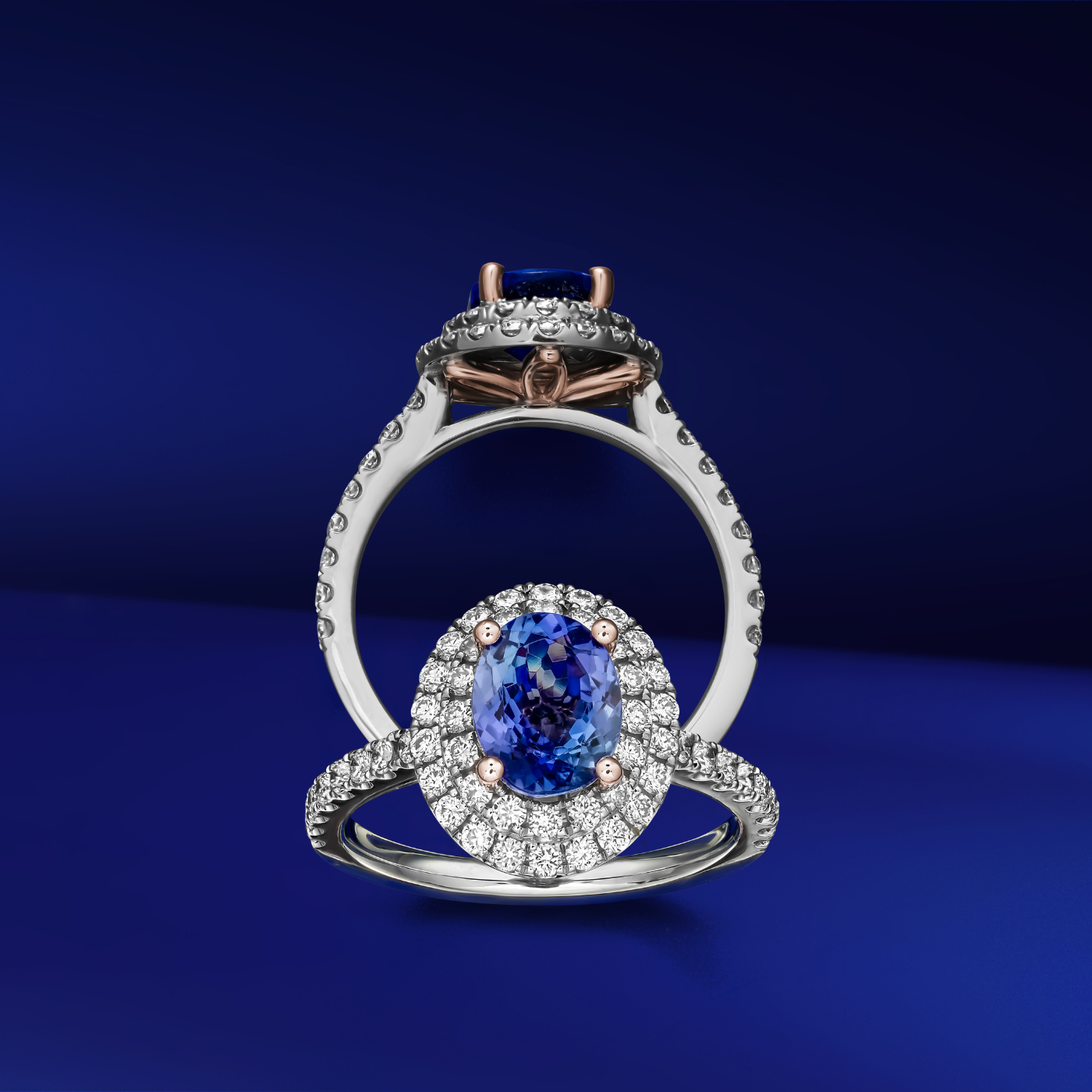 A two row two tone halo ring centered with a oval-shaped tanzanite stone and surrounded by smaller diamonds set in a silver or white gold band from carati jewelry montreal canada white gold and pink gold on blue background