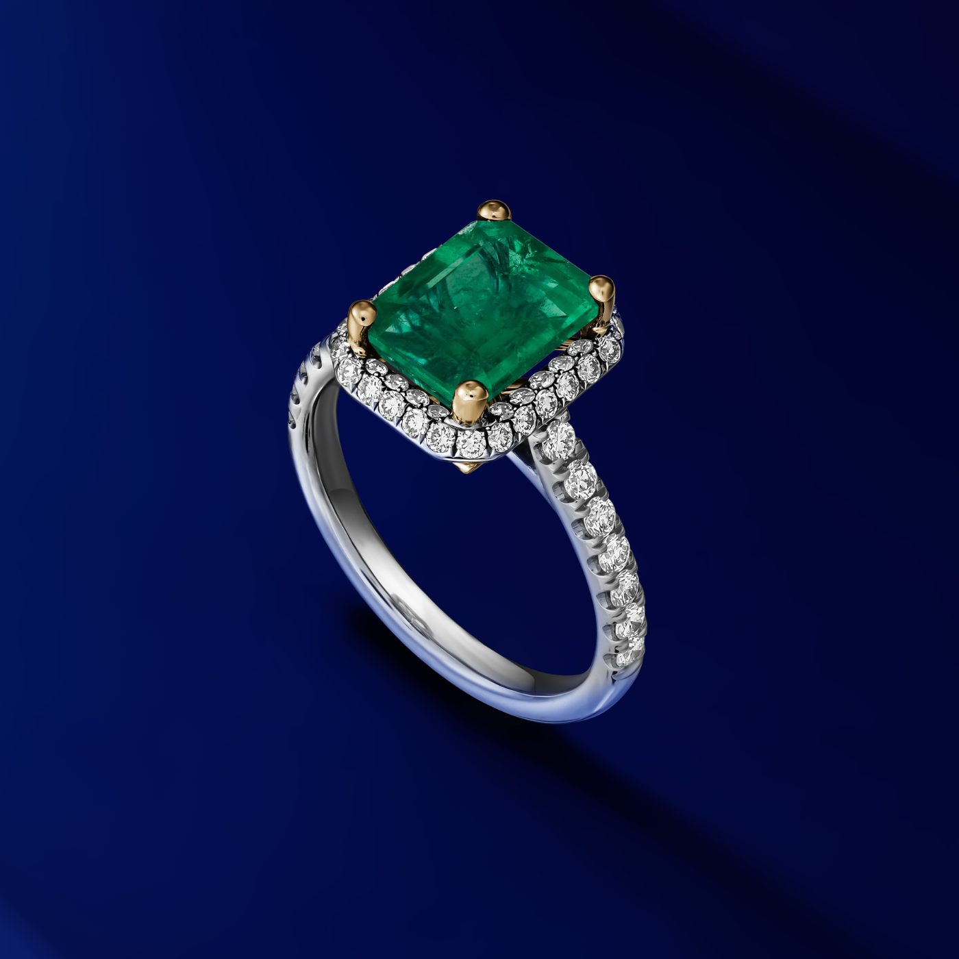 A two-tone gold ring featuring a radiant cut emerald gemstone with a halo of pave set diamonds around it designed by carati jewelry montreal canada 18 k 14 k in a blue backround