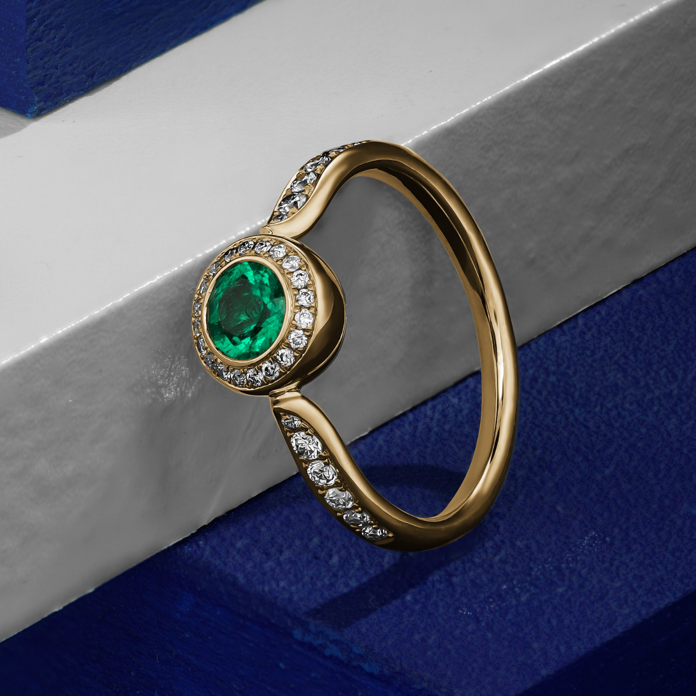 Royal harmony ring with round emerald surrounded by pave diamonds and on the shank designed by carati jewelry in montreal canada 18k 14 k in blue backround