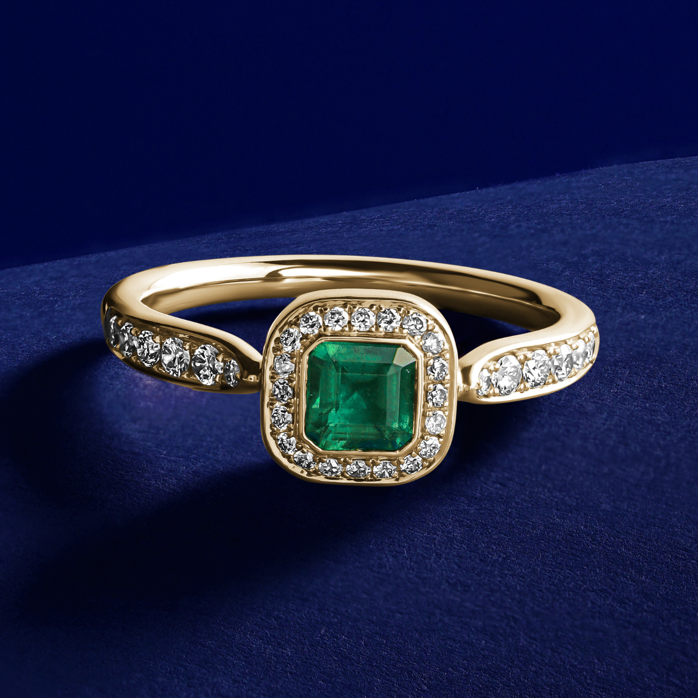 Royal harmony ring with tie emerald surrounded by pave diamonds and on the shank designed by carati jewelry in montreal canada 18k 14 k in blue backround