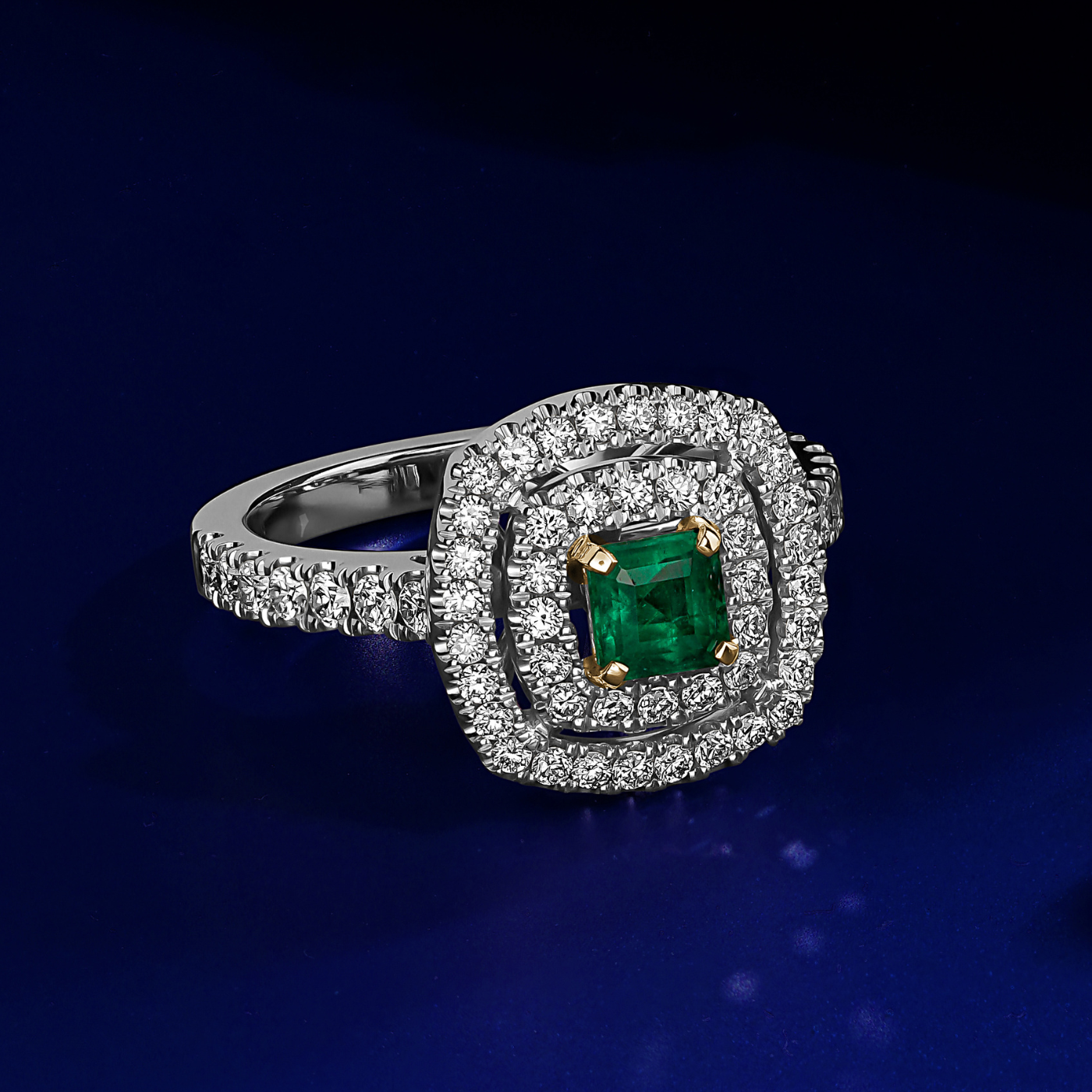 in blue backround A square emerald set in two tone in a double halo ring with a setted shank designed by carati jewelry in 14k 18 k in vs diamonds