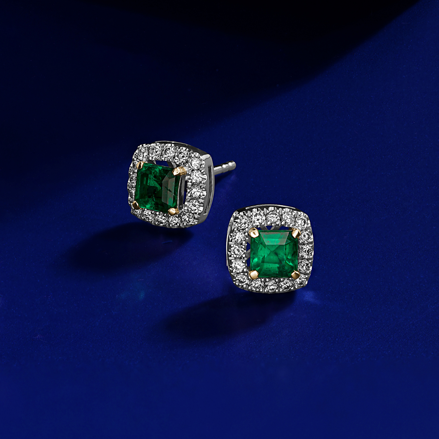 In blue backround A pair of square-shaped emerald earrings with halo sets of sparkling diamonds around each emerald designed by carati jewelry in montreal canada in 18 k 14 k in 2 tone